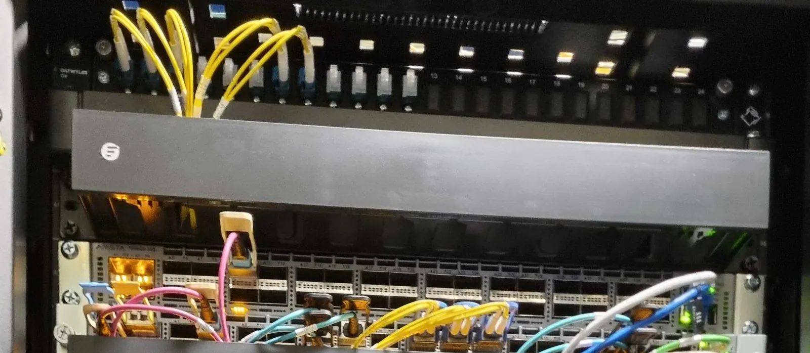 Der obere Teil eines Netzwerk-Racks: ein schwarzes Glasfaserpanel mit gelben Singlemode-Patchkabeln in den linken Ports, eine Kabelführung und darunter ein Arista-Switch mit rosa, blauer und gelber Glasfaser.