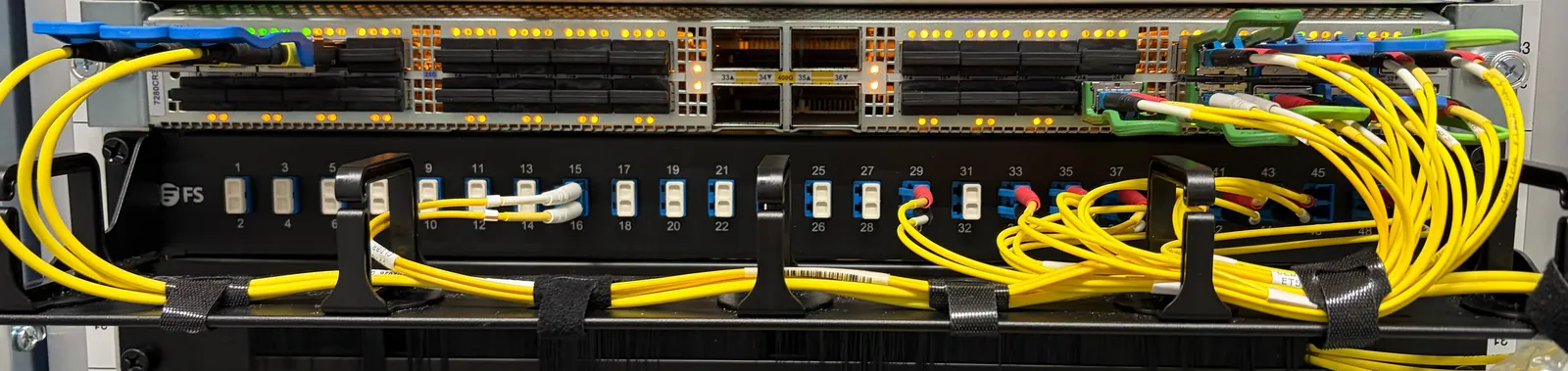Ein Arista-Switch mit bernsteinfarbenen Port-LEDs über einem schwarzen Glasfaser-Patchpanel von FS mit nummerierten Ports, verbunden durch Bündel gelber Singlemode-Glasfaser in Kabelführungen.